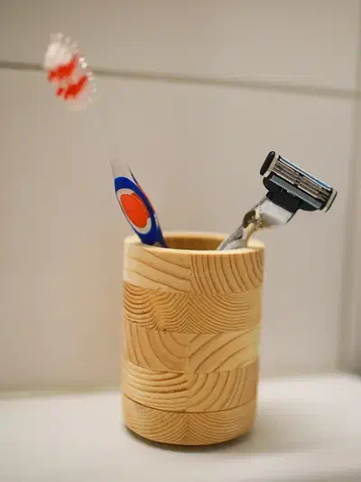 A complete layered wooden cup holds a toothbrush and razor against a tiled wall, with its opening and base unobscured.