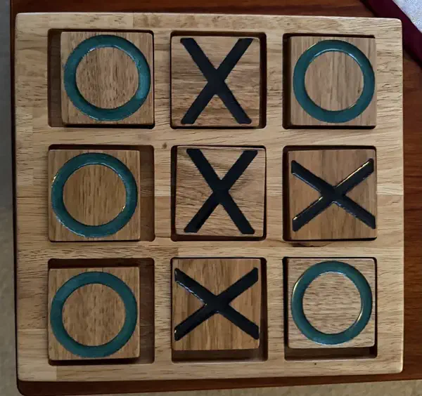 A glossy maple tic-tac-toe board with teal O and dark X grooves filled with colored resin