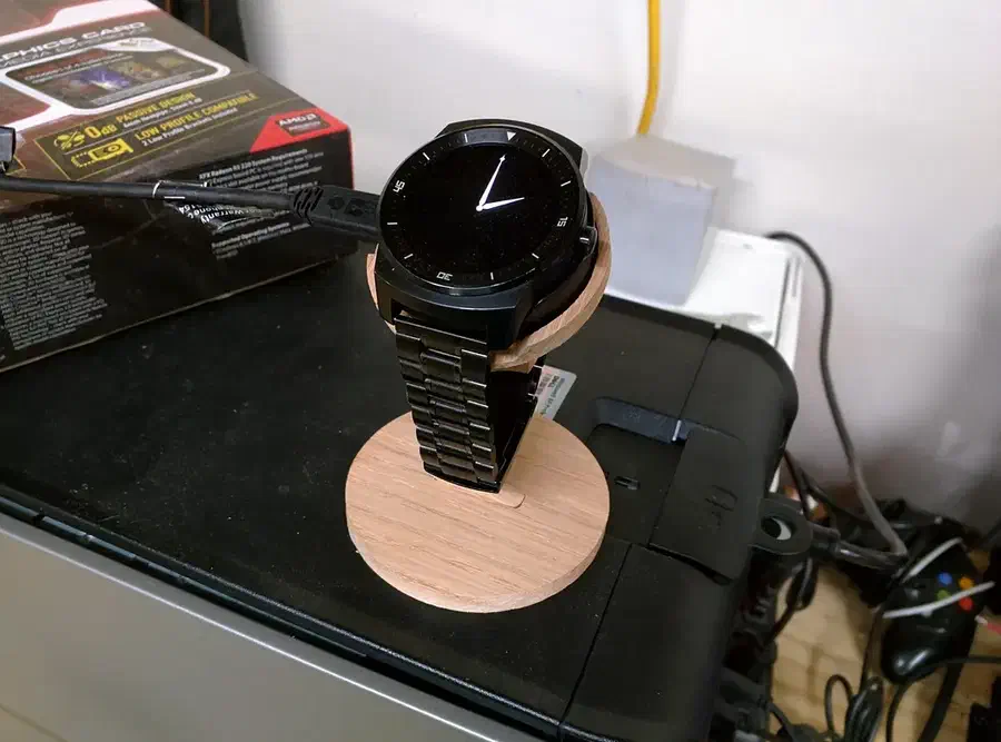 A black round smartwatch on a narrow plywood stand with a round base beside its packaging