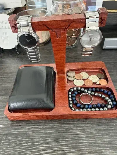 A reddish wooden watch stand with two metal-band watches, a wallet and coins in the base tray