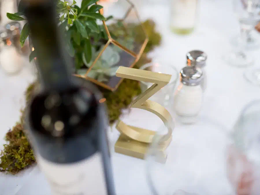 Finished cream-colored standing number three on a wedding table beside greenery and glassware