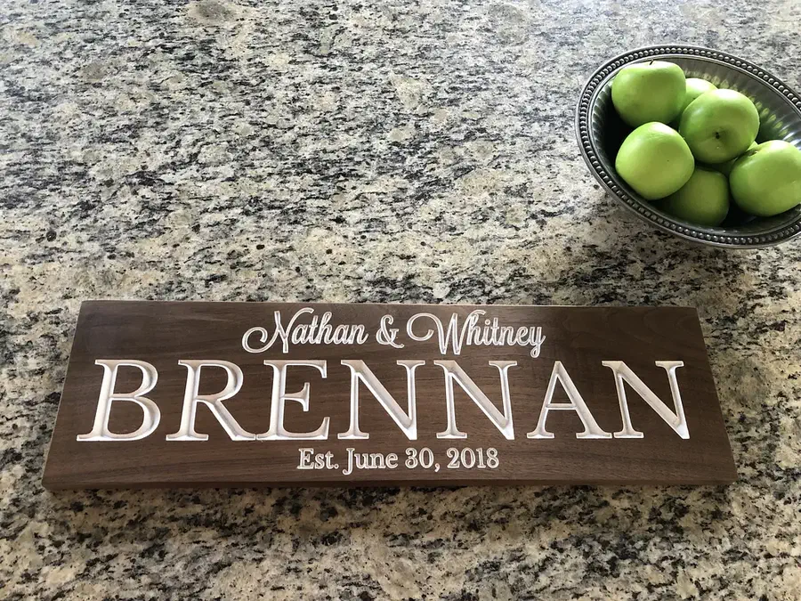 Finished rectangular dark-walnut wedding sign with white carved names, surname, and date on a granite counter